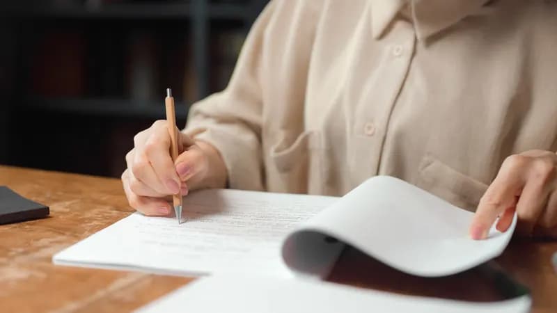 امرأة تجلس إلى مكتب خشبي وتراجع أوراقاً مطبوعة وهي تمسك قلماً وتقلب إحدى الصفحات.
