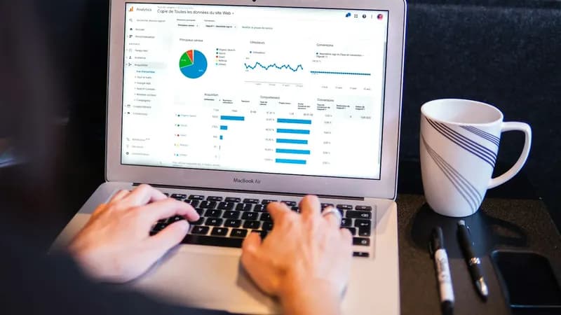 يدان على حاسوب محمول يعرض لوحة تقارير تحليلات لموقع إلكتروني، وبجانبه فنجان وقلمان على مكتب داكن.